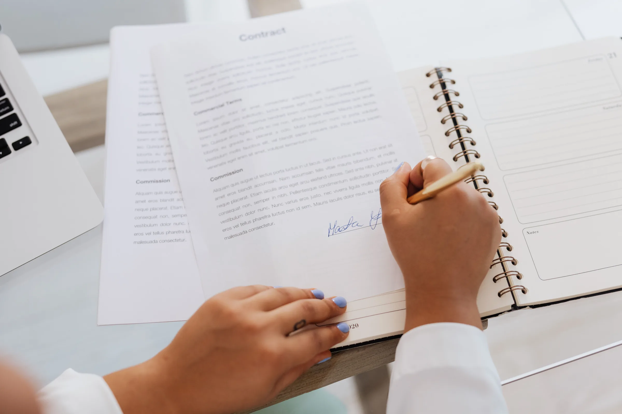 Close-up of hands signing a contract beside a notebook and laptop